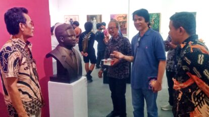 Group of men in a gallery looking at a dark bust on a white pedestal against a pink wall as they chat nearby.