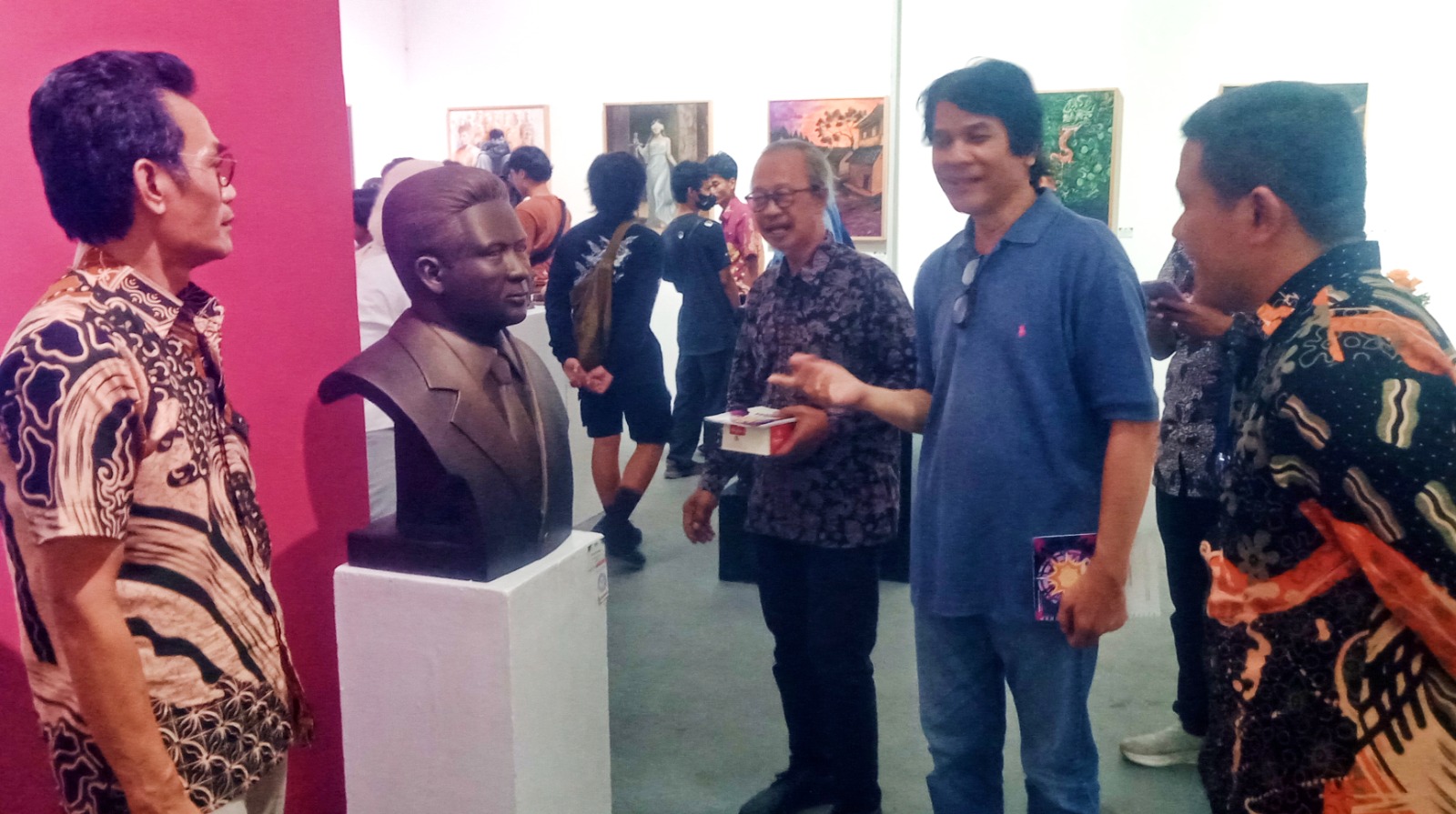 Group of men in a gallery looking at a dark bust on a white pedestal against a pink wall as they chat nearby