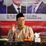 Speaker wearing a black peci hat holding a yellow microphone at a panel with floral arrangement and oranges on the table and a banner with portraits behind him