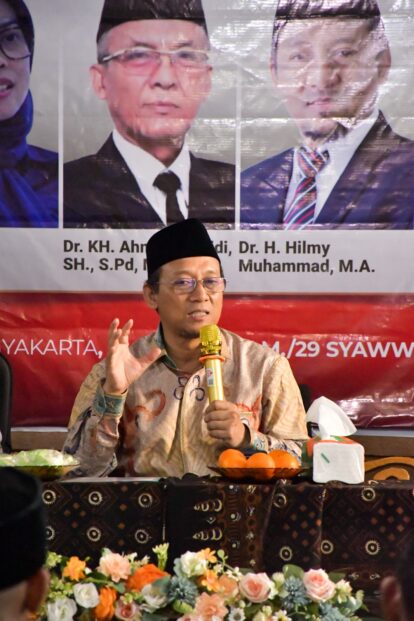 Speaker wearing a black peci hat holding a yellow microphone at a panel, with floral arrangement and oranges on the table and a banner with portraits behind him