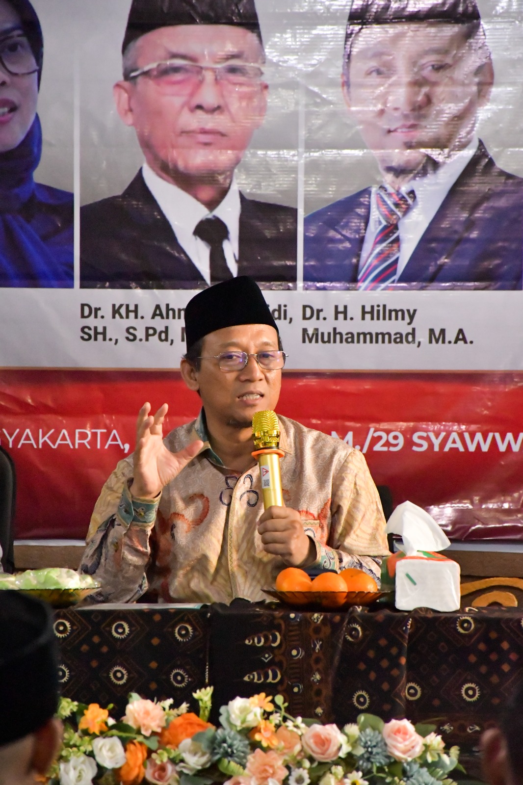Speaker wearing a black peci hat holding a yellow microphone at a panel with floral arrangement and oranges on the table and a banner with portraits behind him