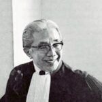 Smiling judge in black robes and white bib sitting in a courtroom wearing glasses