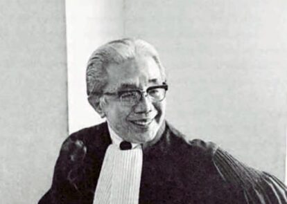 Smiling judge in black robes and white bib sitting in a courtroom, wearing glasses.