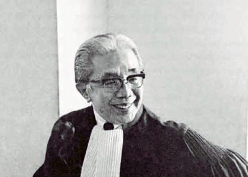 Smiling judge in black robes and white bib sitting in a courtroom wearing glasses