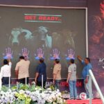 Six men touch a large screen displaying cow silhouettes and blue handprints under a get ready  banner with trophies visible on the left