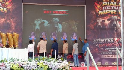 Six men touch a large screen displaying cow silhouettes and blue handprints under a get ready  banner with trophies visible on the left