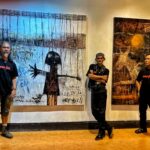 Three men in black shirts stand in front of two large abstract paintings in a gallery setting