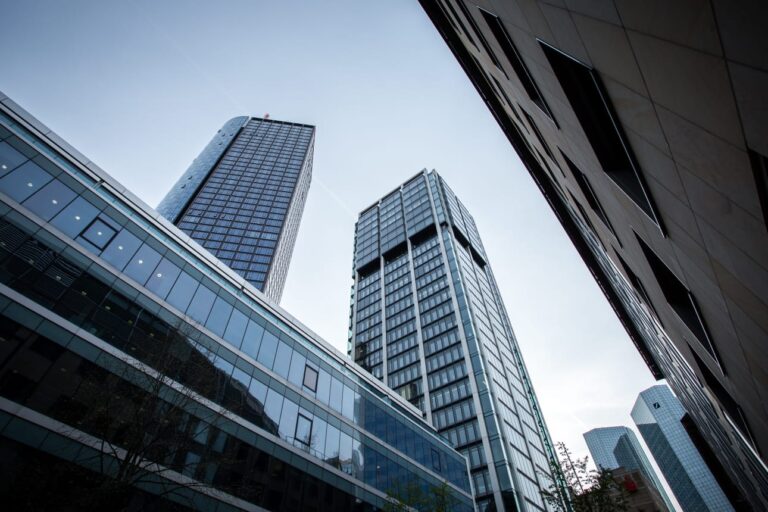 Low-angle view of modern glass skyscrapers against a clear blue sky between surrounding buildings.1/2