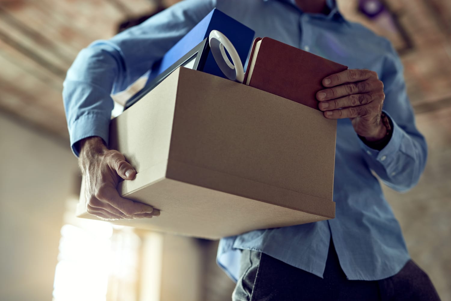 Person wearing a blue shirt carries a cardboard box filled with office items blue binder red notebook mug