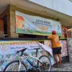 Person in an orange shirt tapes a banner for a daycare program on a storefront with a blue bicycle parked in front and colorful posters along a fence