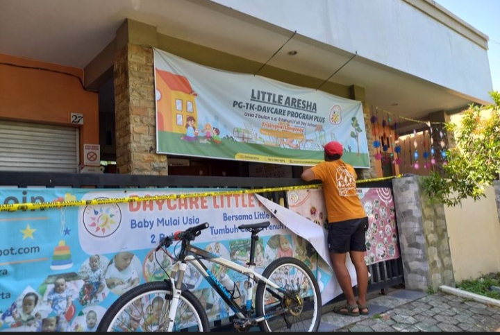 Person in an orange shirt tapes a banner for a daycare program on a storefront with a blue bicycle parked in front and colorful posters along a fence
