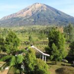 Tall mountain rising over a green valley with trees a red roofed building and a white gazebo in a village garden