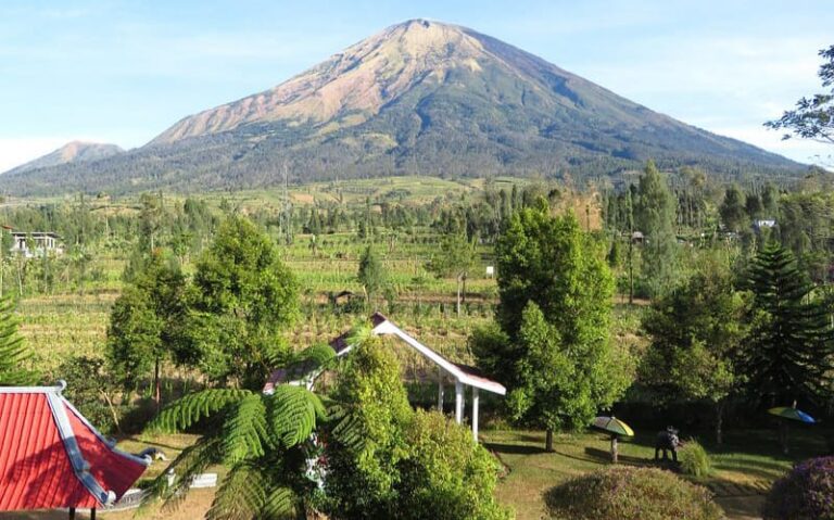 Tall mountain rising over a green valley with trees, a red-roofed building, and a white gazebo in a village garden.