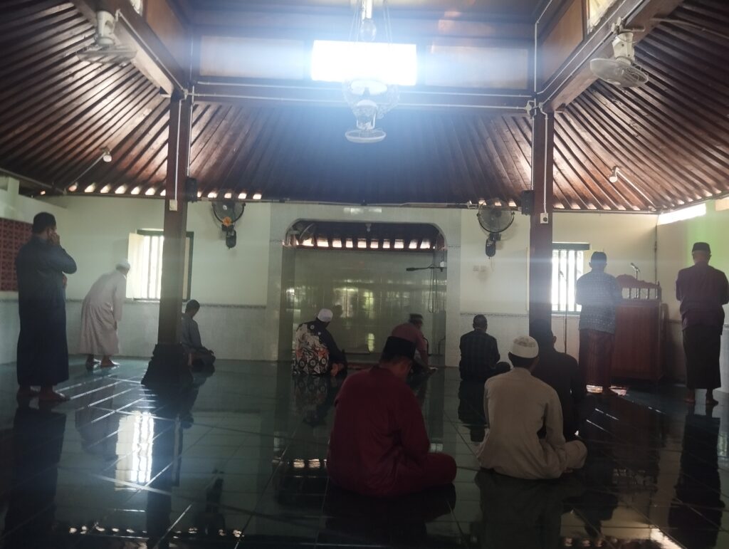 People kneeling in prayer inside a mosque with a wooden vaulted ceiling and fans facing a prayer wallpulpit area on the right
