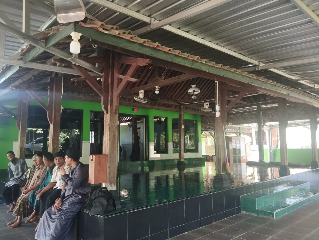 Group of men in traditional clothing seated on a dark tiled bench under a wooden pavilion with a metal roof and green walls