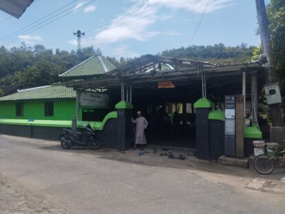 Entrance to a mosque with a green tiled roof; worshippers remove their shoes before entering under a metal-framed shelter.