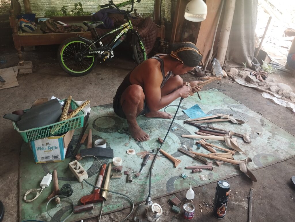 Man kneeling and carving wood on a dusty workshop floor with a neon green bicycle and cluttered tools in the background','Man woodworking in a cluttered shed, surrounded by tools, cords, and a green bicycle nearby.