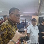Crowd of reporters and officials surround a man in a patterned batik shirt speaking at a press event in a conference room