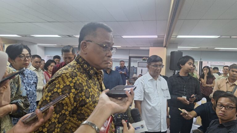 Crowd of reporters and officials surround a man in a patterned batik shirt speaking at a press event in a conference room.