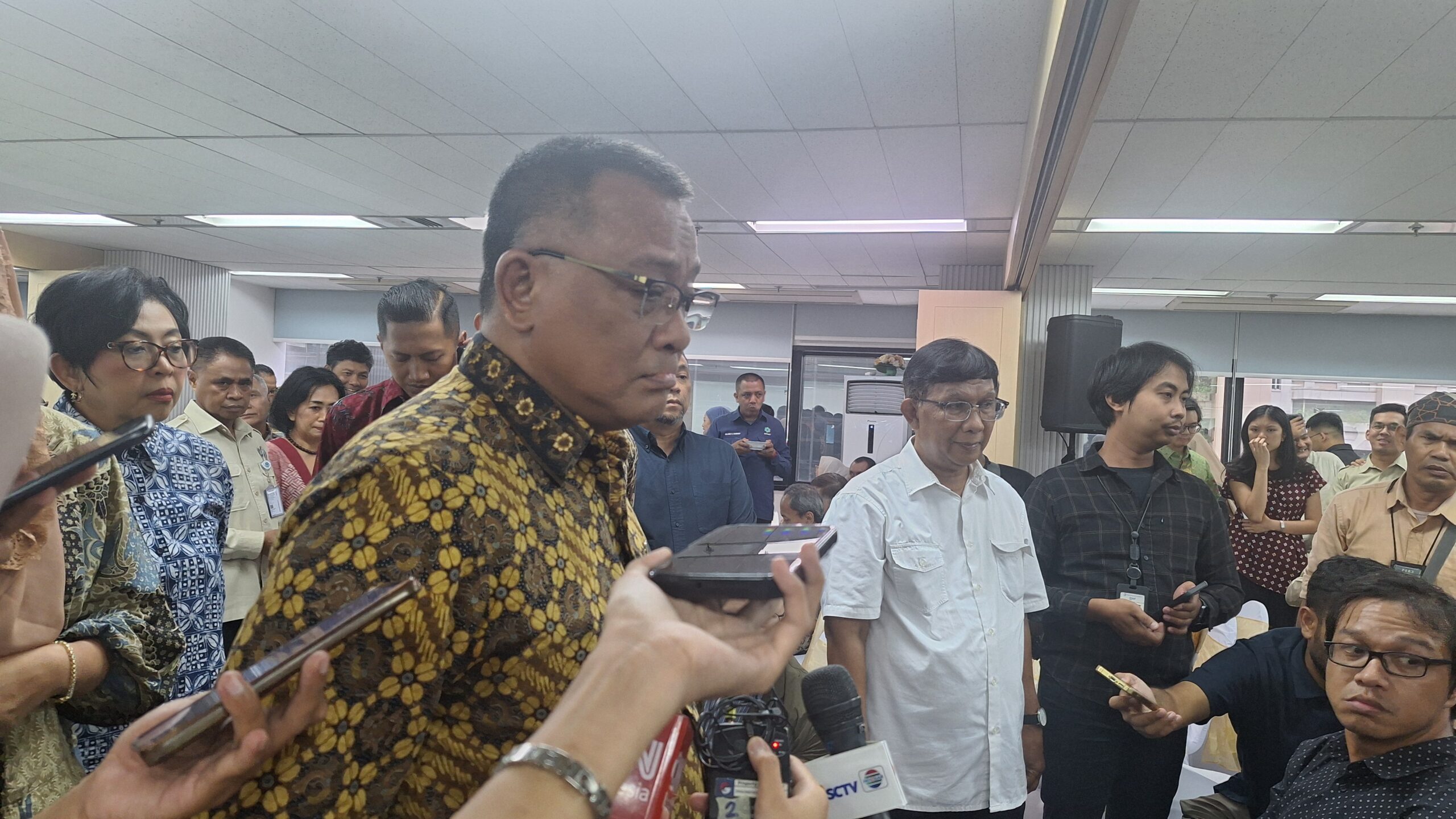 Crowd of reporters and officials surround a man in a patterned batik shirt speaking at a press event in a conference room