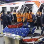 People in orange vests stand near a stopped train on a platform some examining the doorway nearby