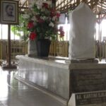 Marble tomb with a draped sculpture on top inside a gated shrine with a vase of red and white flowers nearby