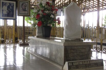 Marble tomb with a draped sculpture on top inside a gated shrine, with a vase of red and white flowers nearby.