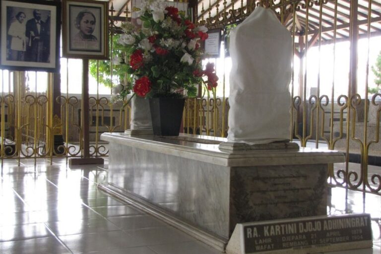 Marble tomb with a draped sculpture on top inside a gated shrine, with a vase of red and white flowers nearby.