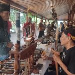 Vendor stall with handmade wooden knives and carved tools on display and two people conversing nearby under a wooden pavilion