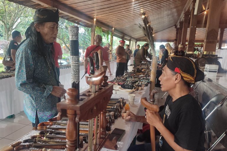 Vendor stall with handmade wooden knives and carved tools on display, and two people conversing nearby under a wooden pavilion