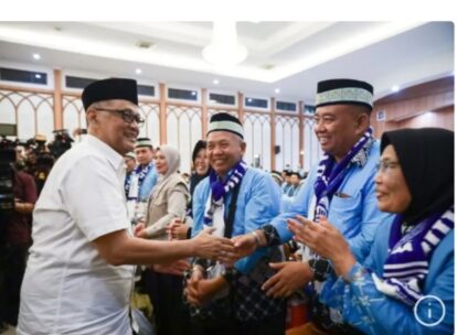 Man in white shirt shakes hands and greets a group of men in blue with scarves at a formal event; smiles all around.
