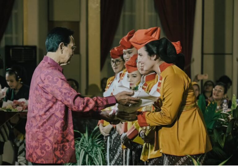Man in a pink patterned shirt presents a bouquet to a line of women in yellow outfits with red headpieces on stage during a ceremony.