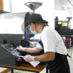Industrial worker wearing mask and cap operates a large coffee roasting machine adjusting controls at a console while another person stands in the background