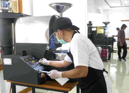 Industrial worker wearing mask and cap operates a large coffee roasting machine, adjusting controls at a console while another person stands in the background.