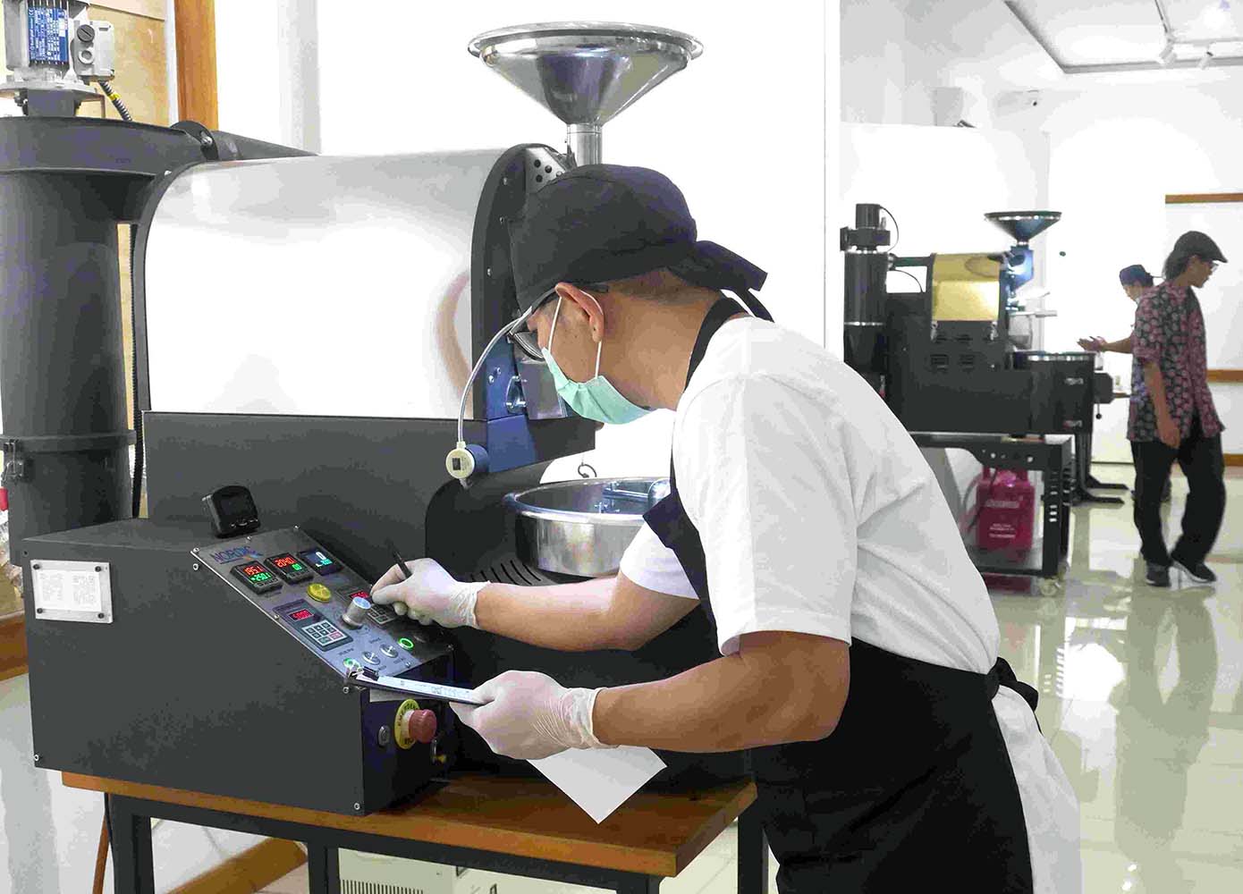 Industrial worker wearing mask and cap operates a large coffee roasting machine adjusting controls at a console while another person stands in the background