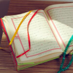 Open arabic book with red border margins and colorful ribbons resting on a wooden surface with turquoise prayer beads draped across it