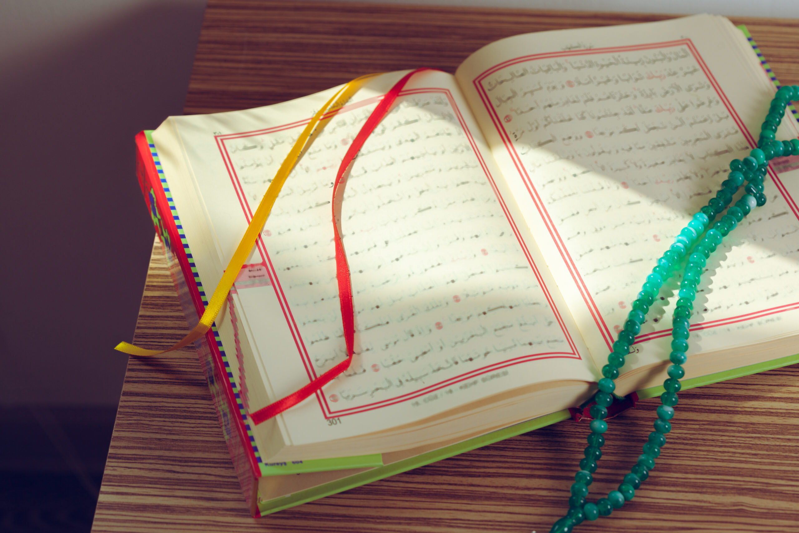 Open arabic book with red border margins and colorful ribbons resting on a wooden surface with turquoise prayer beads draped across it