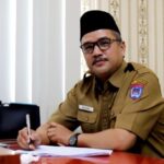 Man in a khaki government uniform sits at a desk and signs a document wearing a name badge and a wristwatch