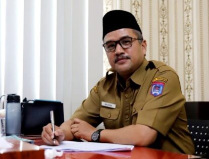 Man in a khaki government uniform sits at a desk and signs a document wearing a name badge and a wristwatch