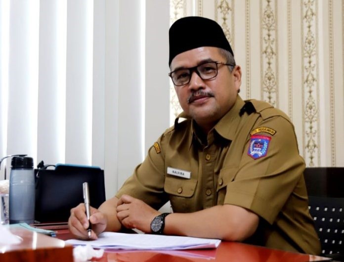 Man in a khaki government uniform sits at a desk and signs a document, wearing a name badge and a wristwatch.