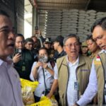 Group of officials in a warehouse with stacked bags a man in a white shirt speaks to the crowd while others wearing badges and vests listen and photograph the scene