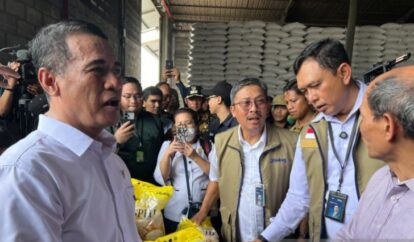 Group of officials in a warehouse with stacked bags a man in a white shirt speaks to the crowd while others wearing badges and vests listen and photograph the scene