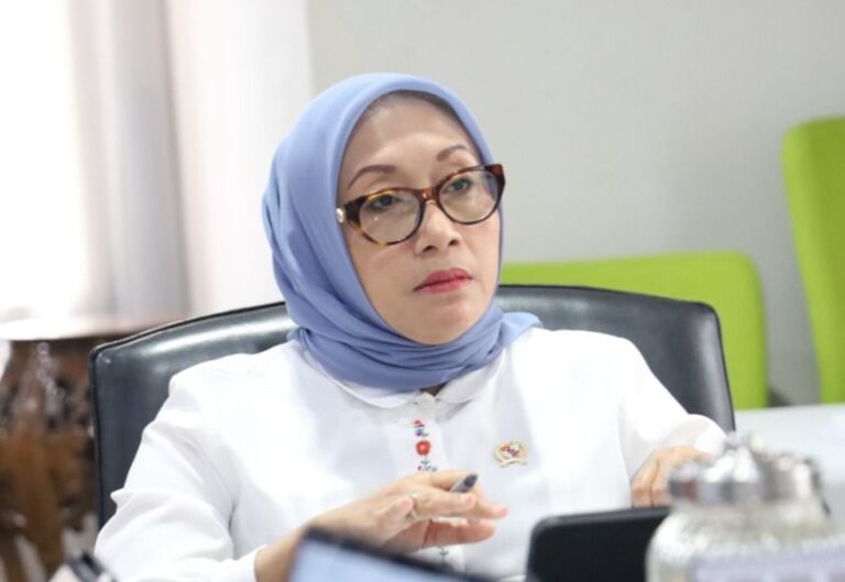 Woman in a light blue hijab and glasses sits at a conference table, looking thoughtful during a meeting.