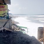 Beachfront shack with a yellow sign reading warung makan mbak tur black sand and bamboo scaffolding by the waters edge