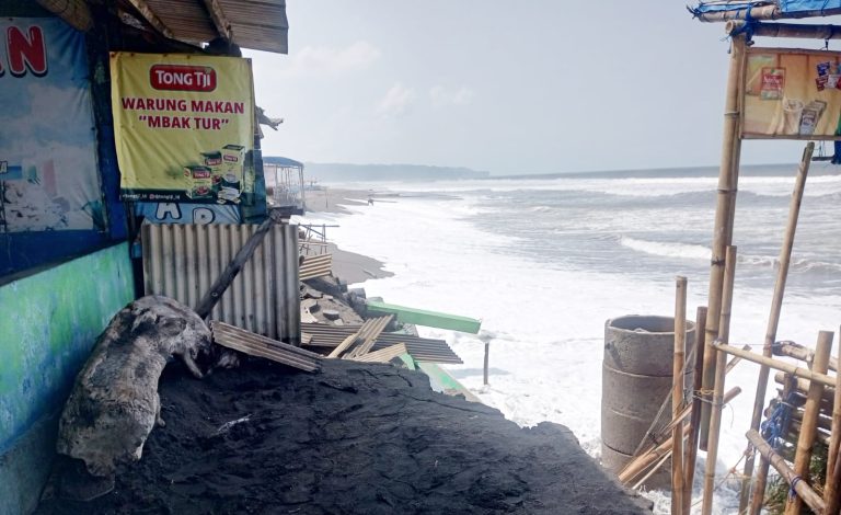Beachfront shack with a yellow sign reading 'WARUNG MAKAN MBak TUR', black sand and bamboo scaffolding by the water's edge.