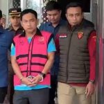 A man in a pink vest with blue shirt and handcuffs is escorted by uniformed officers out of a building