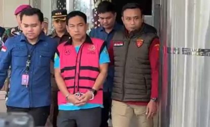 A man in a pink vest with blue shirt and handcuffs is escorted by uniformed officers out of a building.