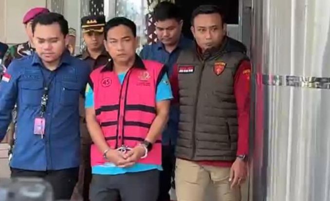 A man in a pink vest with blue shirt and handcuffs is escorted by uniformed officers out of a building