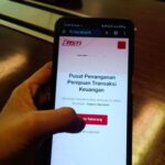 Hand holding a smartphone displaying an indonesian financial fraud reporting page with a red lapor sekarang button on a dark desk