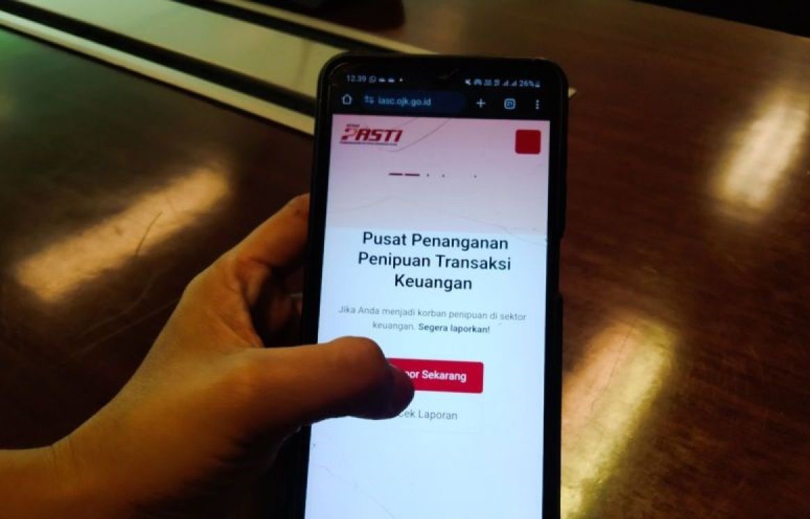 Hand holding a smartphone displaying an indonesian financial fraud reporting page with a red lapor sekarang button on a dark desk